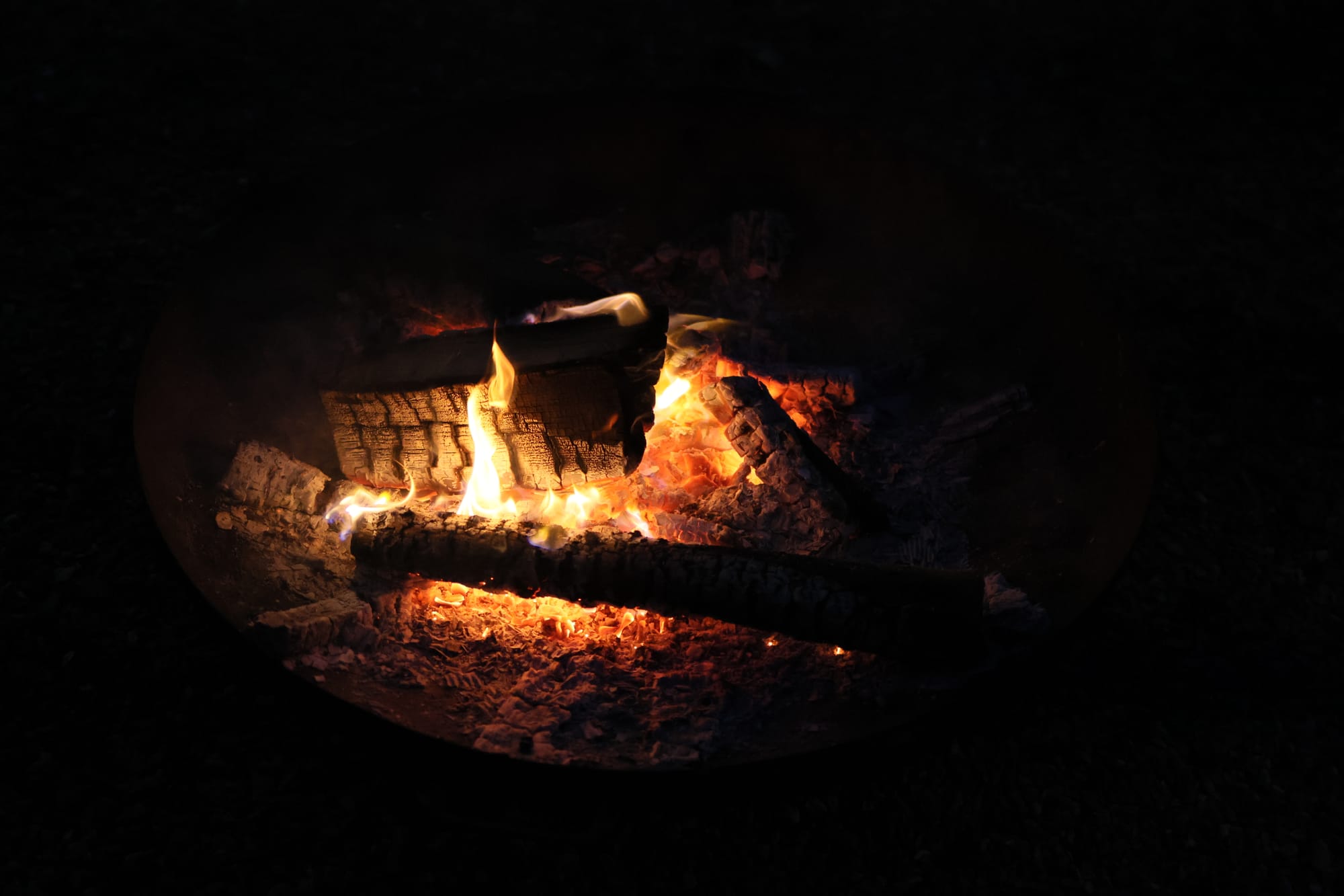 A firepit, full of embers, charred wood and flames licking around a log.